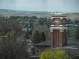 A brick clock tower with white clock faces and black Roman Numerals in front of rolling fields.