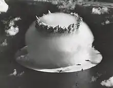 Aerial photo of nuclear explosion rising from lagoon. Hemispherical condensation cloud on the surface is 1 mile (1.6&nbsp;km) in diameter. In comparison, Navy ships in the foreground look like bathtub toys.