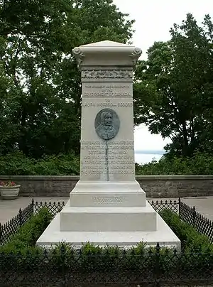 An upright stone monument surrounded by a low hedge.  In the middle is an upright oval of an elderly woman's face in relief.