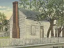 Colorized photo of a log-cabin-style building with a brick chimney and a small tree growing near the door