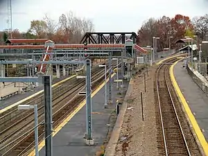 A railway station complex seen from a bridge. At left is a three-track line at ground level; at right is a single-track line rising on an embankment. Both lines have asphalt platforms. In the background, a footbridge and railway bridge over the ground-level line are visible.