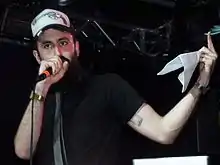 image of a white, bearded rapper singing onstage in a "trucker cap" that has a gun on it