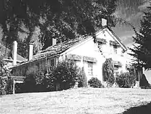 A modest two-story farmhouse with a peaked roof and four chimneys sits under a large tree at the top of a sloping lawn. Bushes and shrubs grow near the house on its two visible sides. A small part of a neighboring house, perhaps only 15 feet (4.6&nbsp;m) away, can be seen in the background.