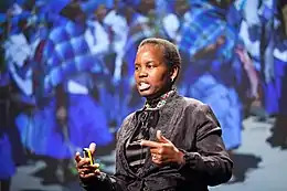 Photograph of an African woman in a grey brocaded suit standing in front of a backdrop depicting a group of African women in blue dresses, while giving a speech. She is wearing a geometrically patterned beaded neck collar, has a yellow device in her right hand, and is pointing with her left index finger to the left of the stage.