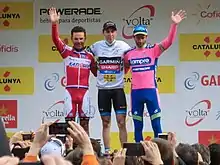 Three riders waving to an assembled crowd with several spectators taking photographs