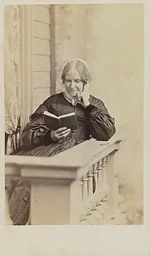  Photographie sépia d'une femme blanche d'âge mûr portant un bonnet blanc, assise et lisant sur un balcon