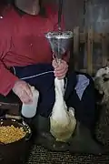 Photo couleur montrant une opératrice assise, tenant coincé entre ses jambes un canard. Un entonnoir est enfoncé dans le bec du volatile et un seau de maïs en grain est posé à côté.