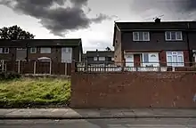 Depuis une rue, vue sur des maisons britanniques du XXe siècle. Les maisons sont surélevées par rapport à la rue. Un terrain vague et herbeux apparaît à la gauche et une mur de briques se trouve à la droite.