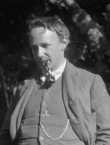 Photographie en noir et blanc d'un homme élégamment vêtu au regard pénétrant, fumant la pipe, une chaîne de montre en évidence sur le gilet de son costume.