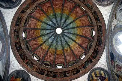 Image photographique de l'intérieur d'une coupole décoré à fresque par des bandes polychromes à teintes se dégradant de la base vers le sommet, les branches d'ogive sont mises en évidence par des teintes plus sombres. Elle comporte à la base seize oculus dont huit sont ouverts alternant avec huit autres contenant des bustes de saints. Au-dessous, le tambour comporte des anges polychromes en bas-relief insérés dans des fausses colonnes à archets et dans les panaches à la base, quatre tondi abritent les Docteurs de l'Église.