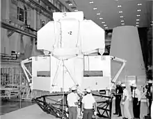 Photographie en noir et blanc d'une maquette du module lunaire LTA-2R en cours d'installation. Des hommes avec des casques blancs discutent devant la maquette.