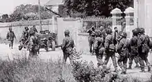 Photo en noir et blanc d'un groupe de soldats marchant sur une route devant le mur d'enceinte d'une propriété.