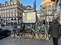 Photographie en couleurs d'une bouche de métro avec portique en fer forgé et des vélos accrochés devant.