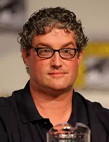 Photographie du buste d'un homme aux cheveux gris bouclés, il porte des lunettes et une chemise noire.