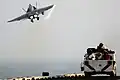 Un F-18 Hornet s'envole derrière un tracteur de pont équipé pour la lutte incendie et monté par des pompiers en tenues argentées.