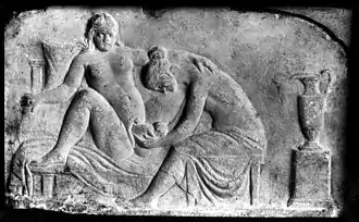 Photographie en noir et blanc d'une scène sculptée permettant de distinguer, sur un fond gris uniforme une femme mettant au monde un enfant, assistée d'une autre femme
