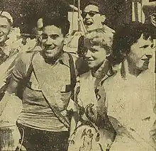 Photo en noir et blanc d'un homme en tenue de cycliste avec une femme blonde à sa gauche. D'autres personnes sont en arrière plan de la photo.