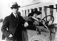 Photographie en noir et blanc d'un homme accoudé à une voiture dans laquelle est assise une femme.