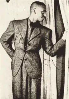 Photographie en noir et blanc d'un homme debout vêtu d'un complet. De sa main gauche il écarte un rideau et regarde ce qui se passe à travers cette ouverture. Son autre main se trouve derrière son dos.