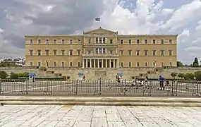 Façade d'un immense bâtiment de pierres jaunes, derrière une vaste place avec des grilles.