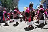 Danse du Pujllay à Tarabuco en 2010. Vue d'ensemble du costume masculin (et féminin en arrière-plan) : on distingue bien les sandales à semelle surélevée, avec leurs énormes éperons parodiques (rappelons que les premiers cavaliers du Nouveau Monde furent les conquistadors), qui servent aussi de sistres pour rythmer le pas.