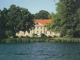 Photo en couleur d'une maison entourée d'arbres, construite en pierres beiges et surmontée d'un toit de tuiles rouges, une étendue d'eau au premier plan.