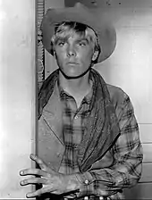 Jeune homme avec un chapeau de cowboy, une chemise à carreaux et une frange derrière un encadrement de porte sur lequel il pose sa main.