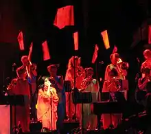 Image d'une chanteuse brune habillée en rouge, entourée par des cuivres et des bagnères