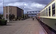 Photographie d'un train arrêté le long d'une plateforme ferroviaire à la fin de laquelle se trouve un pont en arc métallique. Un bâtiment en béton et un panneau portant les insignes de la RDA sont visibles à côté de la plateforme. Plusieurs personnes marchent sur la plateforme et les portes du train sont ouvertes.