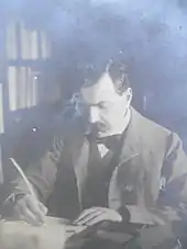 Bourget, assis à son bureau est en train d'écrire, plume dans la main droite, la main gauche posée sur le manuscrit qu'il rédige. Tête baissée et regard concentré. Bibliothèque dans le fond à gauche derrière l'écrivain. Le romancier Paul Bourget.