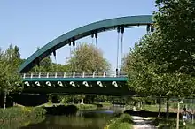  Photo couleurs. Le viaduc de Pannes est vu de côté, sur sa moitié gauche, un arbre au feuillage verdoyant occupe la partie droite de la photo. Le pont est constitué d’un large arc métallique de couleur vert émeraude, sur lequel sont arrimées des barres de fer, appelées suspentes, disposées de manière rayonnante, et qui soutiennent le tablier du pont. Le pont enjambe le canal. Le chemin de halage est à droite de la photo.
