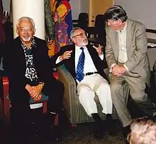 Deux personnages assis sur des fauteuils. un troisième est assis sur un accoudoir et discute avec son occupant.
