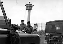 Deux soldats britanniques portant des fusils se tiennent derrière deux jeeps dont l'une porte une plaque «&nbsp;British Frontier Service&nbsp;». Derrière eux se trouve une haute clôture grillagée et une grande tour de surveillance avec un sommet octogonal.