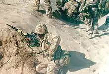 photographie en couleur montrant un soldat accroupi dans une tranchée de sable ; il porte un uniforme camouflé beige et un fusil d’assaut avec un baïonnette fixée au bout du canon