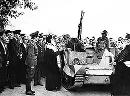 Photographie noir et blanc : un dignitaire orthodoxe bénissant les occupants d'une chenillette, sous les yeux de nombreux hommes, dont des soldats.