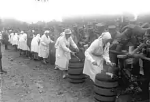 Photo en noir et blanc de sœurs samaritaines en tablier et coiffe blanc, servant à la louche dans la gamelle de soldats placés de l'autre côté d'une barrière, la nourriture qui se trouve dans un tonneau à côté de chacune d'entre elles.
