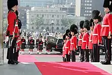 Photo en couleurs de militaires portant une tunique rouge et un bonnet à poil noir et tenant un fusil à la main se tenant debout en deux rangées de chaque côté d'un tapis rouge ; derrière, des militaires porant une tunique verte foncée et un kilt de tenant debout en deux rangées derrière un corbillard dont la porte de derrière est ouverte ; en arrière-plan, des bâtiments d'un centre-ville