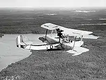 Photo noir et blanc d'un petit hydravion biplan, monomoteur, survolant une zone de forêts et de lacs.