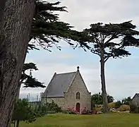 Vue d’une chapelle aux murs de granit et toit en ardoises, avec un arbre en premier plan