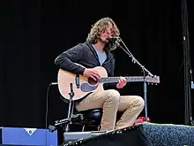 Photographie d'un homme assis sur scène chantant dans un micro et tenant une guitare.