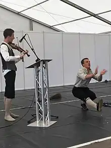 Deux hommes sont sur une scène : l'un joue du violon face à un pupitre, l'autre exécute un numéro de danse.