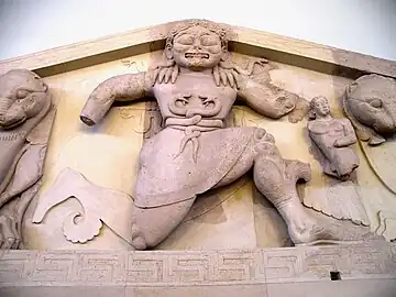 Méduse a des serpents sur les épaules, les bras étendus, le genou droit au sol. La bouche est très large, les yeux sont clos.