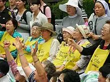 Anciennes femmes de réconfort, aujourd'hui très âgées, alignées et assises lors d'une manifestation, elles arborent des tabliers sur lesquels sont écrites des revendications.
