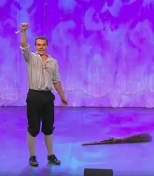 Un homme blanc portant des sabots lève le poing, debout sur une scène éclairée en violet. Un balai est aussi posé au sol.