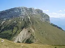 Barres rocheuses dominant une grande pente herbeuse parcourue d'un sentier en zigzag.