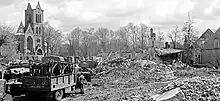 Photographie en noir et blanc montrant les ruines d'une ville, avec les murs d'une église (sans toit) encore visible. Au premier plan, un camion militaire.