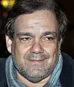 Visage rond d'un homme d'une cinquantaine d'années, mal rasé, arborant un léger sourire.