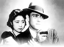 Une femme se tenant aux côtés d'un homme portant un chapeau pointe un revolver (image en noir et blanc).