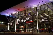 Photo de nuit de l'entrée vitrée éclairée d'un bâtiment d'exposition sur laquelle est suspendue une affiche « Equita’ ».