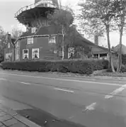 Photographie en noir et blanc d'une maison et d'une remise couronnés d'une galerie et longées par une route.